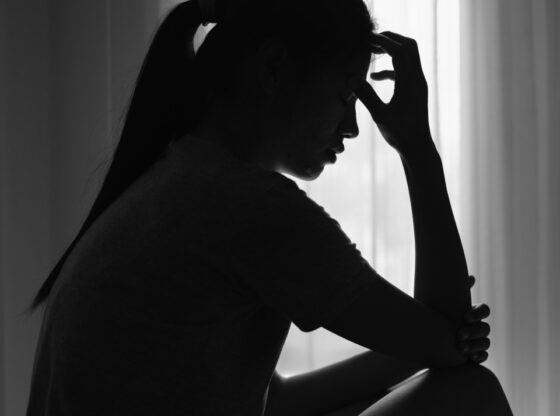 Silhouette of a young woman sitting in a dark room, head down, holding her forehead, depicting pain or distress.