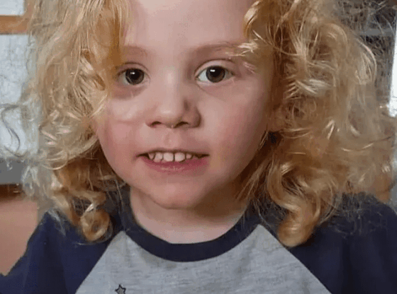 A young child with blonde curly hair smiles slightly at the camera, wearing a gray shirt with the text "MY MUMMY" and a pink pig character.