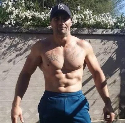 Man wearing a New York Yankees baseball cap and blue shorts, showing his torso with tattoos.