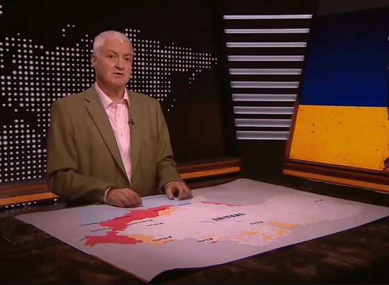 A man presenting on a news set with a map of Ukraine showing areas of conflict, and a torn Ukrainian and Russian flag on the right.