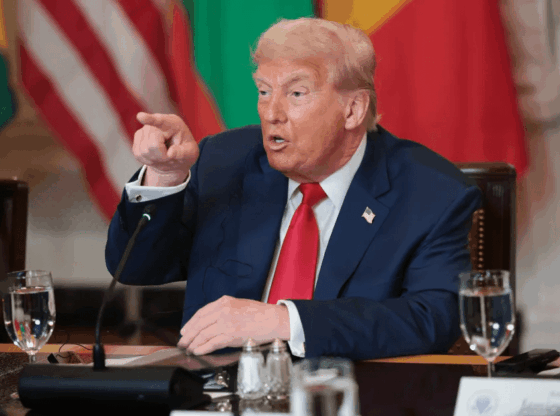 Donald Trump in a blue suit and red tie, sitting at a table with flags in the background, pointing with his right hand.